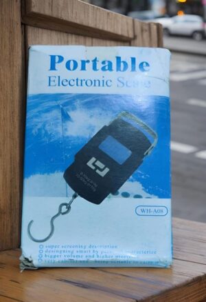 Box for a portable electronic scale showing a handheld scale device with a hook, leaning on a wooden surface beside a street.