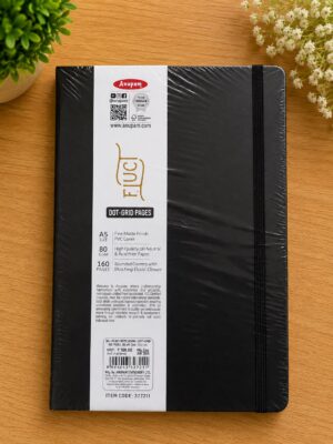 Black notebook wrapped in plastic with a white product label band showing brand, specs, and an elastic strap on a wooden surface with plants nearby.