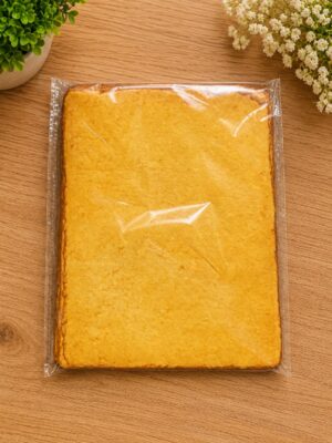 Large rectangular sponge cake wrapped in plastic on a wooden table with green plants nearby.