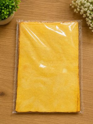 Top-down view of a large rectangular yellow sponge cake wrapped in clear plastic on a wooden table, with green plants nearby.