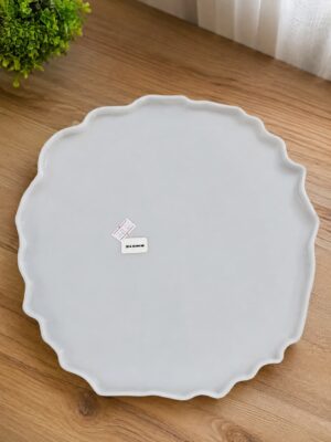 White ceramic plate with a scalloped edge resting on a wooden table, with a small price tag nearby and a green plant in the corner.
