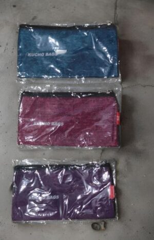 Three plastic-wrapped clutch bags in blue, pink, and purple stacked vertically on a concrete floor (Kucho Bags).
