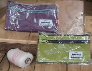 Two striped zipper pouches in plastic wrap on a wooden surface, one purple and one green with labels visible, plus a spool of white thread nearby.