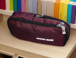 Maroon fabric pencil case with dual zippers on a wooden desk; colorful books lined up behind.