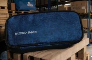 Blue zippered bag with 'KUCHO BAGS' logo resting on a wooden pallet in a warehouse environment.