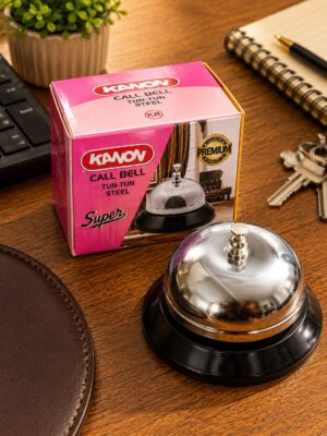 Pink Kanon call bell box on a wooden desk with a shiny chrome service bell in the foreground and office items nearby