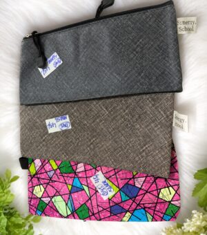 Three zippered pouches stacked diagonally: dark gray textured, light gray textured, and a bright pink geometric-pattern pouch, with small brand tags attached.