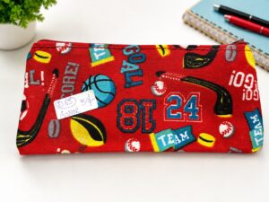 Red sports-themed pencil case with football, basketball, and team graphics; a small handwritten label is attached to the surface.