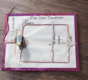 Step-down transformer model mounted on a pink poster board with red wires looping around a hand-drawn circuit diagram labeled 'Step Down Transformer'.