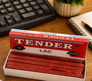 Open box of red licorice sticks labeled 'Tender Lac' on a wooden desk beside a keyboard, notebook, and plant.