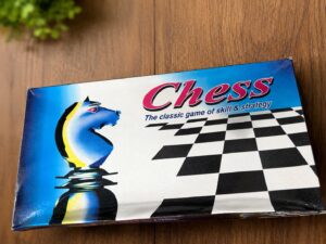 Box for a chess game on a wooden table, featuring a blue knight piece and a black-and-white checkerboard pattern with the word 'Chess' and the subtitle 'The classic game of skill & strategy'