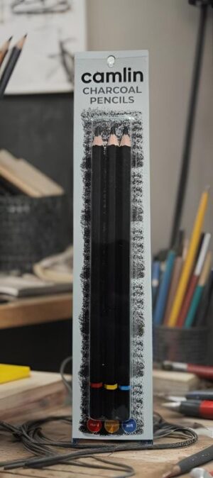 Boxed set of Camlin charcoal pencils with three black pencils and color rings at the bottom, on a cluttered art desk.