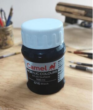 Close-up of a Camel acrylic paint bottle (Black, 016) with a light blue cap on a wooden art desk, brushes in the background.