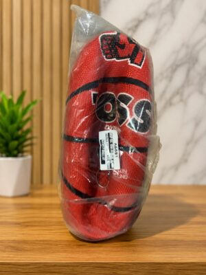 Red packaged product wrapped in clear plastic, standing upright on a wooden table with a barcode sticker visible. Background includes a small plant and a marble wall.