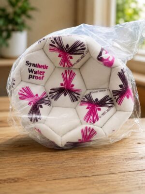 White synthetic waterproof soccer ball with pink/black starburst designs, wrapped in clear plastic on a wooden table.