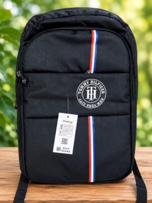 Black Tommy Hilfiger backpack with red, white, and blue stripe, placed on a wooden table with a tag attached.